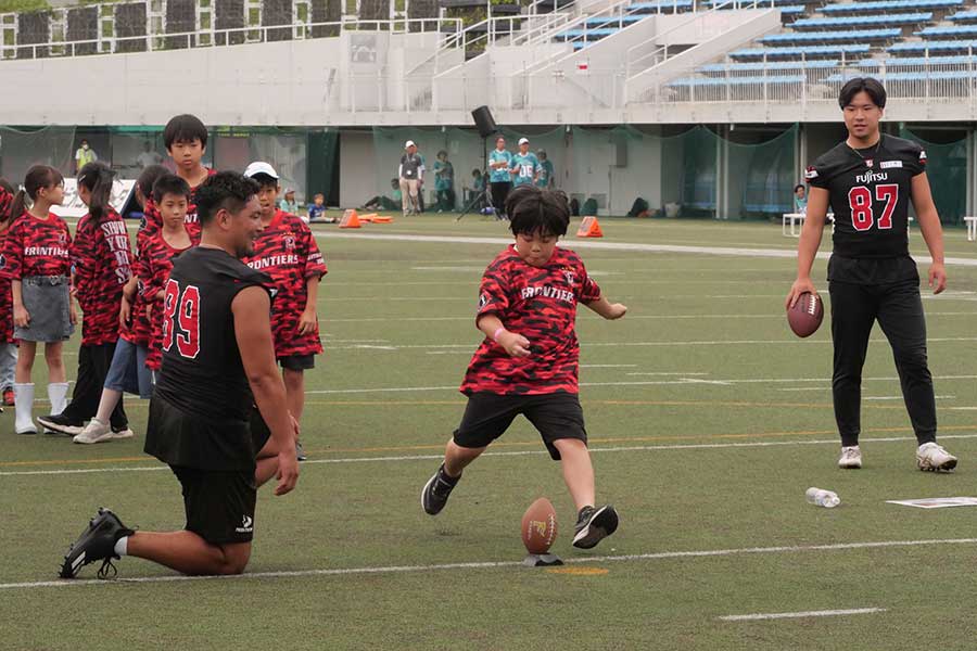 ボールを蹴る参加者の子供(中央)と笑顔で交流を深める富士通アメリカンフットボール部・福井雄哉(左)【写真:上田悠太】